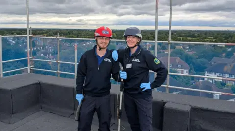RSPCA Ben Fitzcosta and Meg Lloyd wearing dark blue jumpers and trousers, with light blue gloves. Ben is on the left wearing a red helmet, Meg is on the right wearing a dark blue helmet. They are looking into the camera and smiling while stood on the roof of a building. Behind them is scaffolding and a view over houses and trees