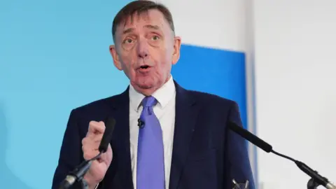 Sir Robin Wales speaks into a microphone at a press conference, in front of a blue backdrop.