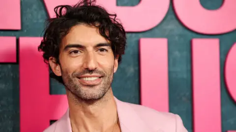 Reuters Justin Baldoni in a pink suit posing in front of a grey backdrop with large pink letters at a film premiere