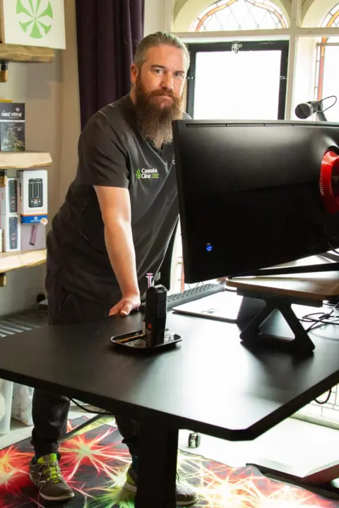 A man in scrubs with a long dark beard and tied back dark hair stand above a black desk in an office. Cannabis posters and medical books line the shelves behind him. 
