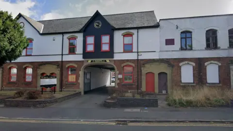 A part brick, part white rendered building, with red window frames. An access arch in the centre, with a clock is above it