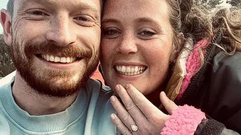Victoria Rodwell A grainy, selfie shows Victoria Rodwell and her partner, Ben Stevens, in a close-up photograph on the day they got engaged. She is holding up her left hand to show her engagement ring. He is wearing a light blue jumper and she has a black jacket with with a pink fluffy scarf underneath. They are both smiling and looking happy.