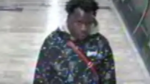 British Transport Police A blurred CCTV image  of a man walking in a corridor of Oxford Circus tube station. He has black hair and is wearing a patterned jacket and has a red strap across his chest. 