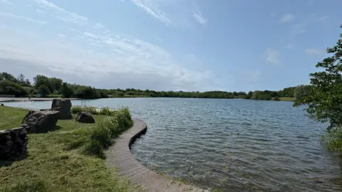 A general view of the Cosmeston Lakes Country Park. The lake is in the background and to the left of the picture there is a path, a grassy verge with some rocks on it.