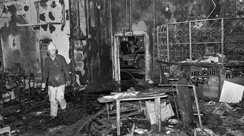 Independent News And Media/Getty Images A black and white photo of a firefighter in the burned out wreckage of the Stardust nightclub in Dublin in 1981. He is wearing a bright-coloured helmet and a protective uniform. The roof, walls and floor of the building have all sustained extensive fire and smoke damage. The remains of damaged tables and chairs are strewn around the floor. 