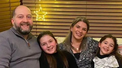 Ellie Goan A family photo including a mother, father and two daughters sitting on a couch inside what looks to be a living room. The mother who has long dark hair is sitting in the middle, she is wearing black nylons and a silver top. Her young daughters who are sitting either side of hair also have dark hair. The father has some grey hair and a beard, is wearing a grey top and trousers and is sitting to the left of frame. There are star shaped light decorations on the wooden Venetian blinds behind the couch.