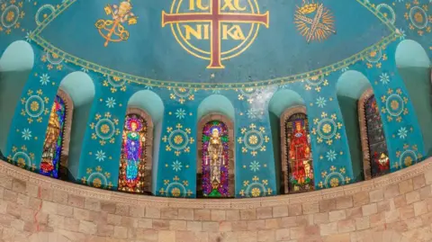 Historic England Three stained glass windows in a kaleidoscope of colours, featuring Catholic saints.