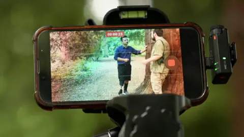 The friends stand by a tree talking which is captured in the image in an iphone on a stand.
