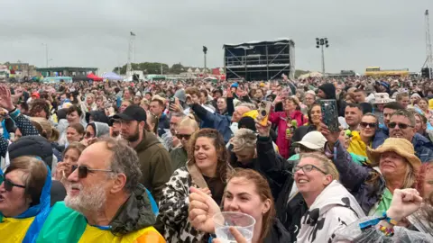 Image of a happy-looking crowd of people stretching far into the background. Festival tannoys and stalls can be seen in the far distance.
