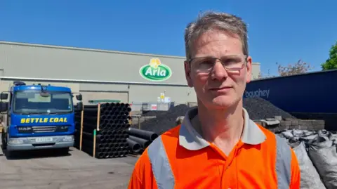 A man with short grey hair and glasses looks at the camera. He is wearing a hi-vis orange polo shirt. Behind him is a pile of coal, a lorry with the words "Settle Coal" on it, and a building with the Arla logo on the side.