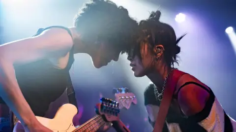 Two female guitar players touch heads on stage during a performance.