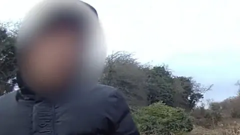 A blurred our face of a boy wearing a padded black top in front of some trees.