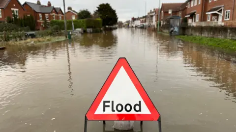 A triangle warning sign saying 'flood' in middle. The sign has been placed in flood water on a road in front of homes.