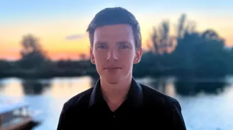 Become A white man with short black hair. He is looking at the camera and wearing a black shirt. He is standing in front of a lake. Behind the lake are black silhouettes of trees and a yellow and orange sunset