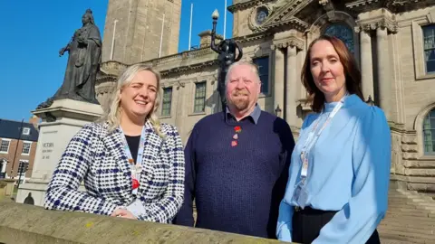 South Tyneside Council Standing in front of South Shields town hall are Laura Turner, who has blonde hair and wears a black and white checked jacket, Paul Dean, who has a short grey goatee beard and short grey hair, dressed in a blue jumper, and Lisa Murphy, who has brown hair and wears a smart light blue blouse. A statue of Queen Victoria is in the background.