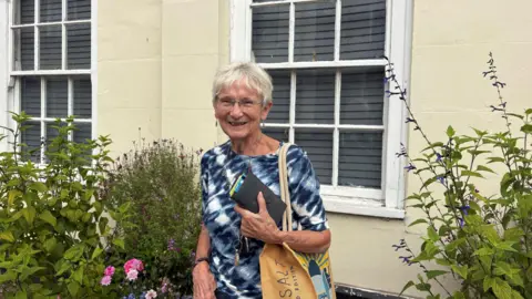 A smiling older lady wearing a dark blue dress with white patterning and short cropped white hair stands outside a cream-coloured building