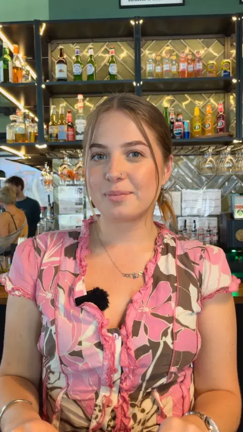 Person seated at a bar or café table, facing the camera, wearing a pink floral-patterned top with ruffle trim and a small microphone clipped to the neckline. Behind the person, illuminated shelves display bottles of beer, spirits, and soft drinks, with hanging glassware and a tiled backsplash creating a warm, modern bar setting.