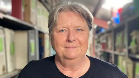 Sarah Ingleby is stood in a storage room with lots of machines on racking to her left and right. She has short grey hair and is wearing a black T-shirt.