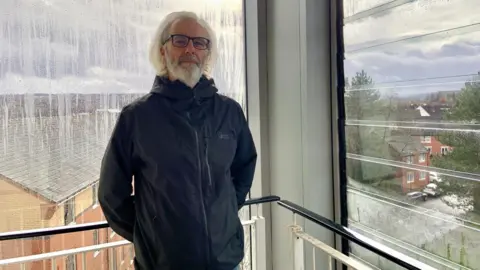 A man with shoulder length grey hair and a grey beard is wearing glasses and a black jacket. He is standing on the corridor of an apartment block by a window showing the street below
