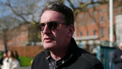 PA Media A man wearing a black jacket and sunglasses. He is standing outside with red brock buildings in the background.