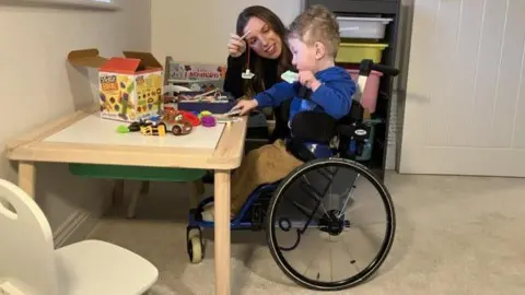 A young boy with curly blond hair and wearing a blue top and beige trousers is sat in a wheelchair at a small table. A woman with long dark hair in a dark top is sat next to him and they are playing together with toys on the table.