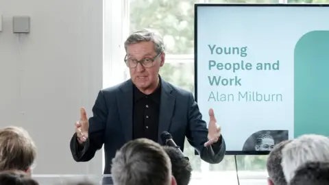 Jeff Moore/PA A man with short grey hair wearing a grey suit and black shirt addresses a room full of people. He is wearing round glasses and gesturing with his arms