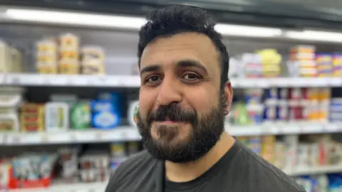 A headshot of Can Duran. Items in fridges can been seen in the background.