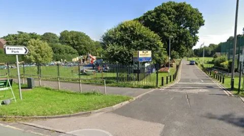 Google The entrance to a park. A signpost saying Ravenhill Park can be seen on the left, and a long driveway with a single car approaching. A children's playpark is on teh left and a sign for Brereton Town FC. There is a further grass area on the right.