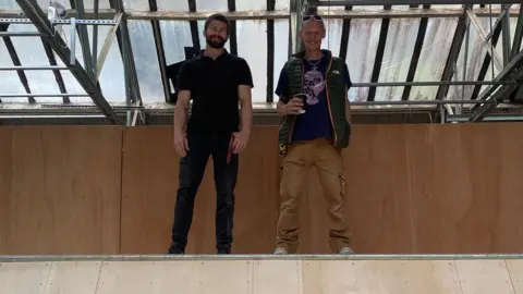 BSS Two men stand at the top of a wooden skateboard ramp, looking down at the camera. The man on the left has short dark hair and a dark beard and is wearing jeans and a dark polo shirt. The man on the right is wearing brown trousers and a blue t-shirt under a green sleeveless jacket. He is holding a takeaway coffee cup and has a pair of sunglasses perched on his head.