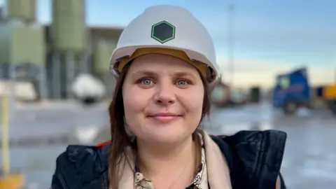 Material Evolution CEO, Dr Liz Gilligan, wearing a hard hat and standing outside the Wrexham factory. Part of the factory and a blue lorry cab can be seen behind her