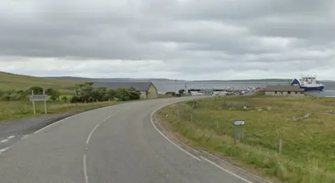 The road near Toft ferry terminal. The roads runs through an area of fields. There is a blue and white ferry at the terminal. It is a grey, cloudy day.