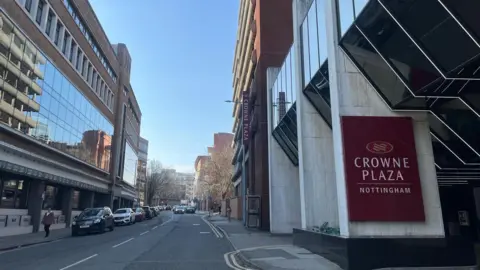 A photograph of Wollaton Street in Nottingham, on the right is the Crown Plaza hotel, which is tall. A red sign for the hotel can been seen on a white wall. On the other side of the road there is another tall building 