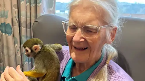 Ruth Cox has long silver hair tied up in a ponytail and is holding a squirrel monkey which is looking at the camera. She is sat in a chat and wearing a pink cardigan and green polo shirt.