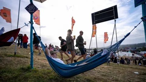 PA Media A man in a white T-shirt is lying in a blue hammock which is secured atop a grassy hill. People in the background are walking across the hill and a white festival tent can be seen further down.