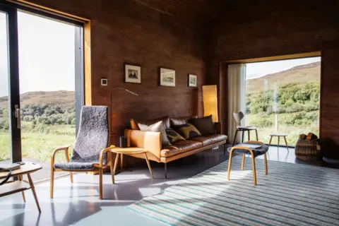 IWC Media A warm, wood-panelled living room features a leather sofa, minimalist chairs, and a striped rug arranged in a clean, modern layout.
Large floor-to-ceiling windows frame a sunlit rural landscape, blending the cosy interior with the surrounding greenery.