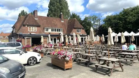LDRS Several tables with benches are outside the front of a pub, which appears to date from around the 1930s. Cars are parked on the edge of the beer garden.