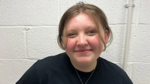 Oliver Conopo/ BBC A woman with her brown hair tied back is looking at the camera smiling. She is wearing a black t shirt and is standing in front of a white wall. 