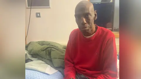 Cherelle Thompson Clive sits looking sad wearing a red jumper and sitting on a hospital bed. He is older and thinner than in his picture from his youth and wears a hospital wristband.