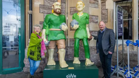 GWR A woman and man standing either side of the Avon Gorge Giants statue outside of Temple Meads Station. They are both smiling at the camera and holding onto it.