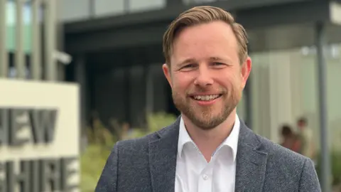 Head and shoulders image of Alex Beckett. He has brunette hair swept to the side, a beard and is wearing a navy suit and white shirt.