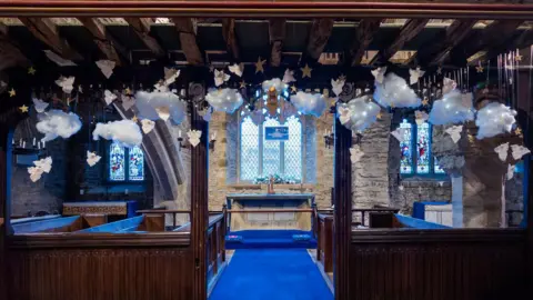 Sandra Kilvington Interior of a historic stone church adorned with decorations. Above the altar, cotton-like clouds, knitted angels, and golden stars hang from wooden beams.