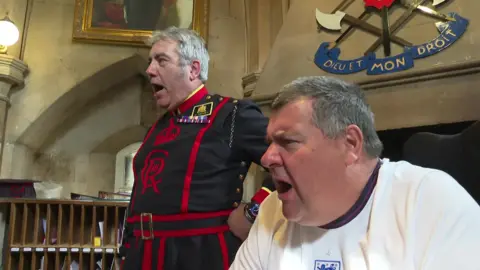 The two men in the historic room watch the game and react with a shout. A fireplace with a crest bearing the words “Dieu et Mon Droit” is visible behind them.