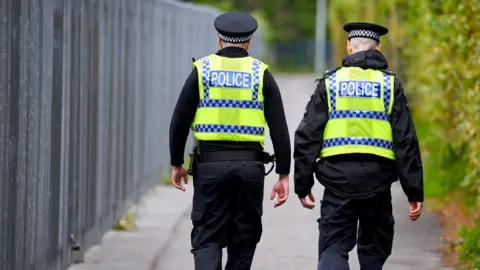 Getty Images Two police officers are walking down a street. They are both wearing hi-vis vests over a black uniform and wearing black hats. There are bushes to their right and a metal fence to their left.