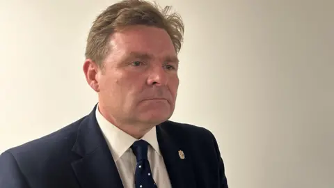 BBC Birmingham City Council leader John Cotton, wearing a navy blue suit with a pin badge on the lapel. He is also wearing a navy coloured tie with white spots.