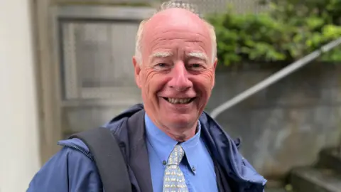 Deputy George Oswald - A man with a bald head and white hair on the side of his head. He's wearing a blue coat, blue shirt and a pastel blue tie. 