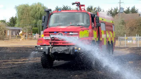 BBC A picture of a red fire truck being driven to spray water out of a hose on its front.