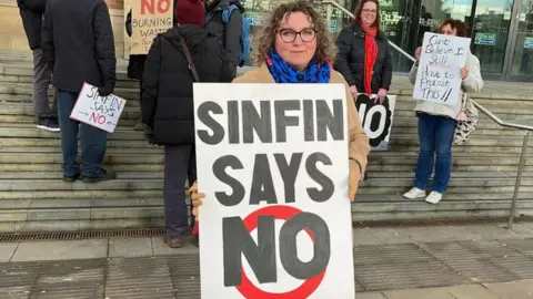 Woman stands on stone steps of Derby City Council house infront of other protesters holding large signs that say "Sinfin says no"