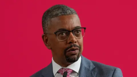 Vaughan Gething wearing a grey suit, white shirt and a red flowery tie. He is wearing glasses with black rims at the top and is standing in front of a red background.