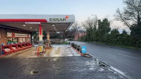 There is a petrol station with Essar branding with four bays. There is red open boxes on the far left with white signage. On the right-hand side, there is a road with a line of trees to its right. 