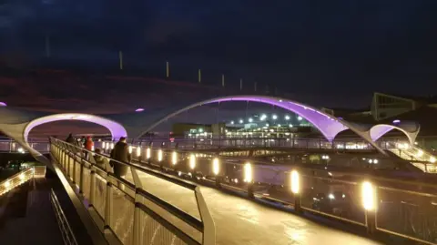 A bridge with arches lit up underneath with purple lighting over a road. The picture is taken at night.